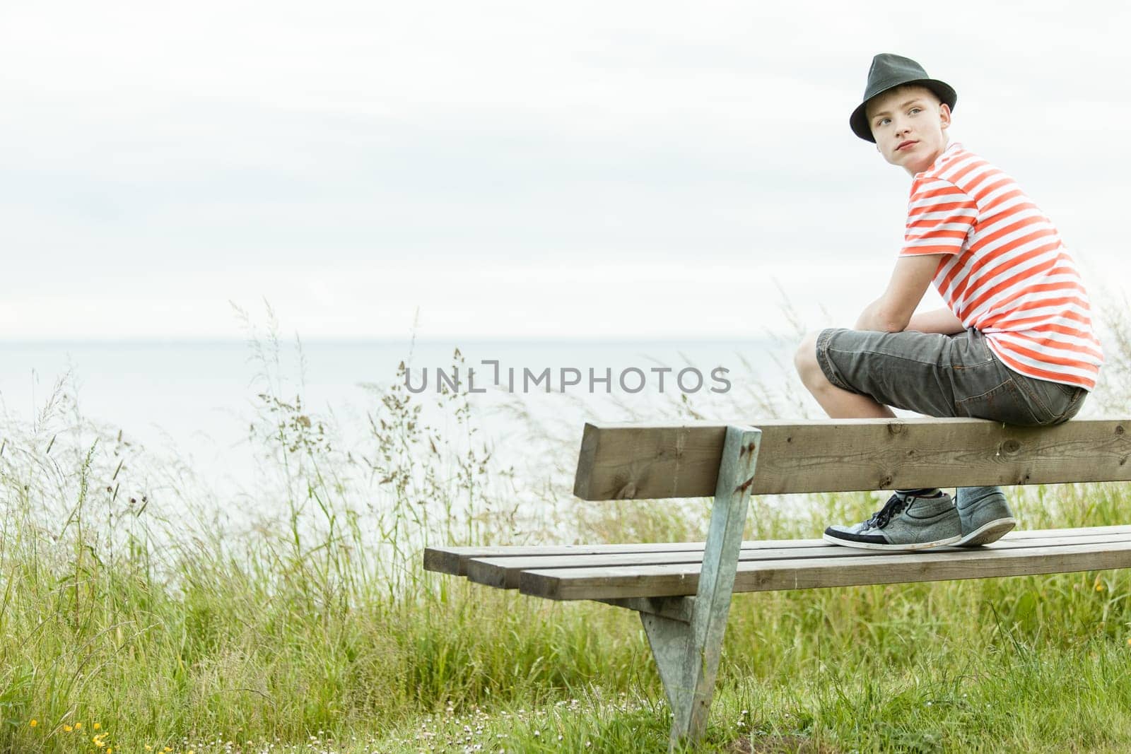 Rear view of teenager in striped shirt and hat sitting on top of wooden bench in front of ocean looking over his shoulder
