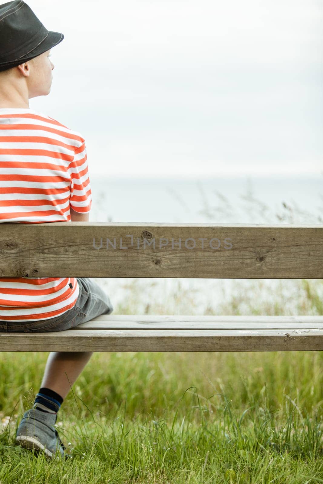 Rear view of single teen in black hat and striped shirt sitting on one side of bench facing grass and ocean