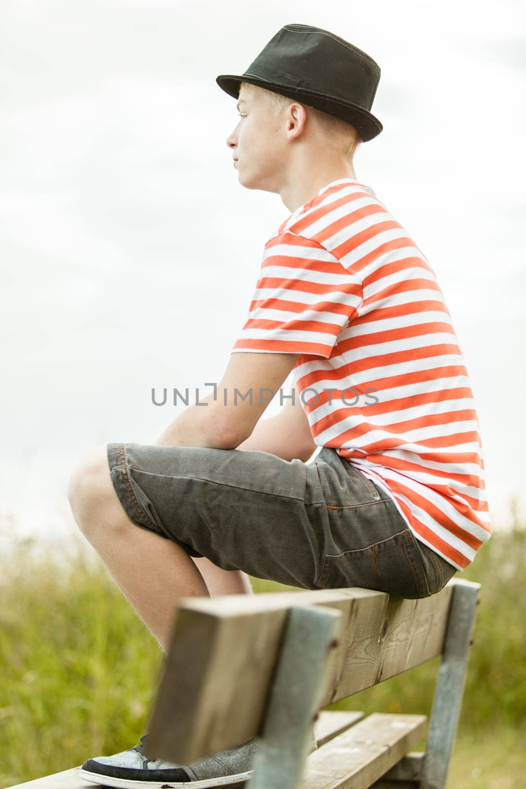 Side view of calm male teenager in hat, striped shirt and shorts sitting on top of wooden bench near wild grass