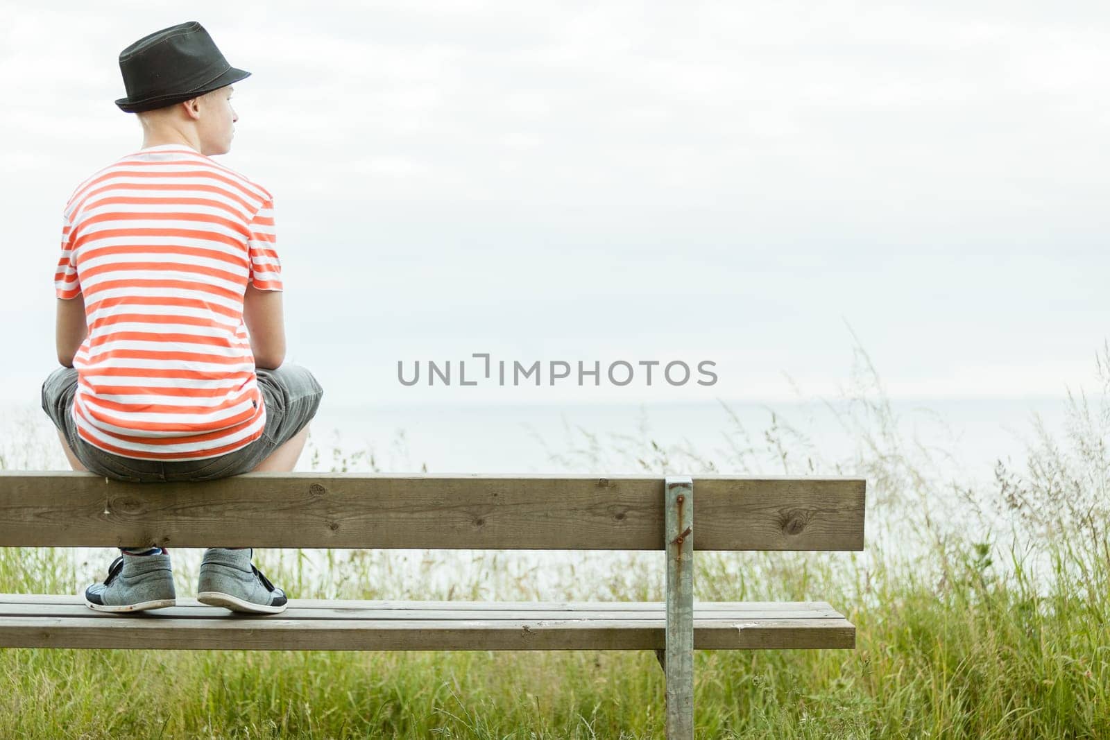Teenage boy in a trendy hat sitting on a rustic wooden bench overlooking the sea with his back to the camera and copy space