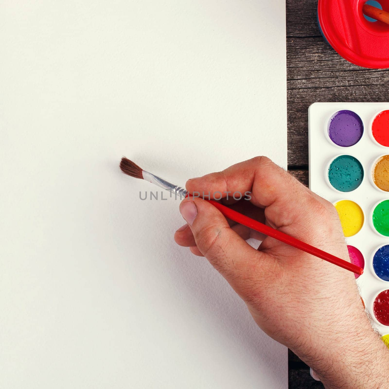 hand with brush painting with watercolor  and blank white paper sheet  on vintage wooden background