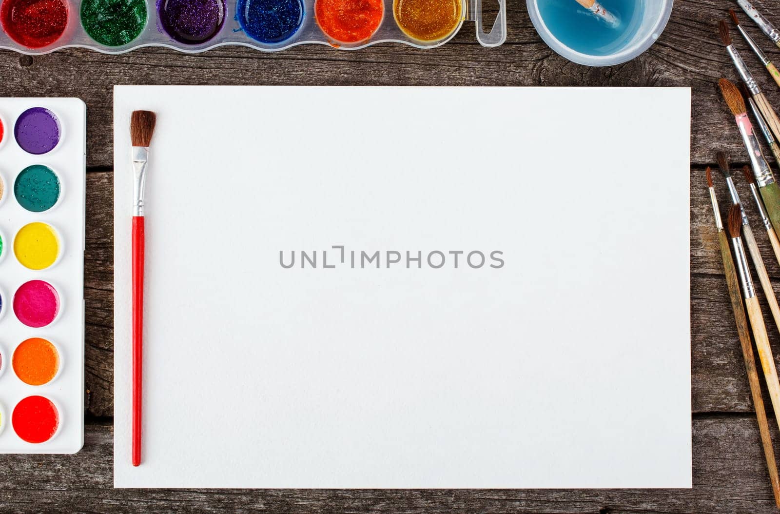 Set of watercolor paints,  brushes for painting and blank white paper sheet of sketchbook on vintage wooden background. Top view.