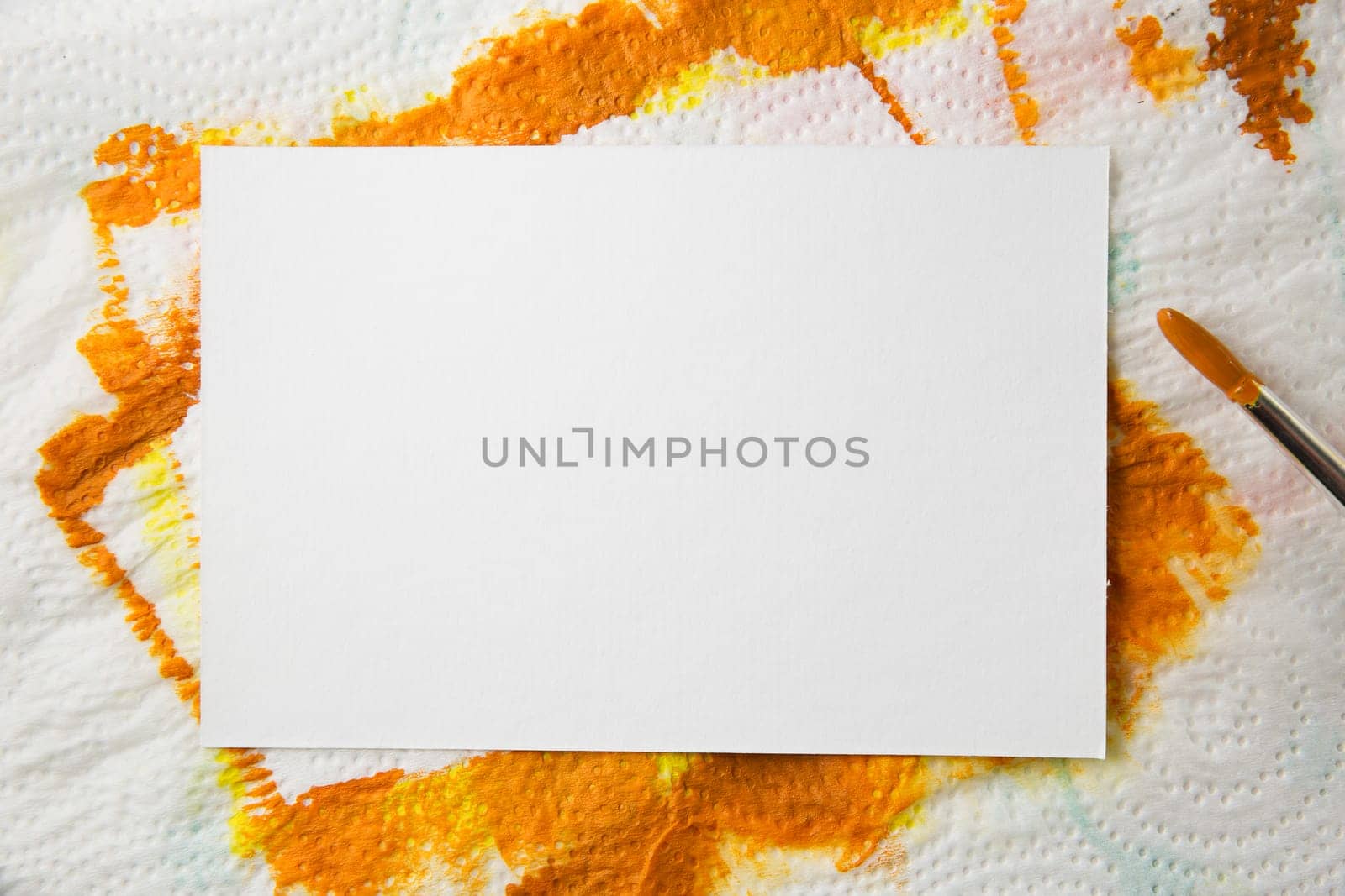 Blank white watercolor paper on towel with orange paint and paintbursh covered with orange paint.