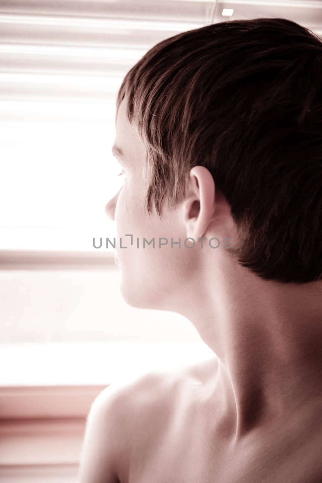 Close up head shot of a shirtless thoughtful young boy staring out of a bright window viewed in profile