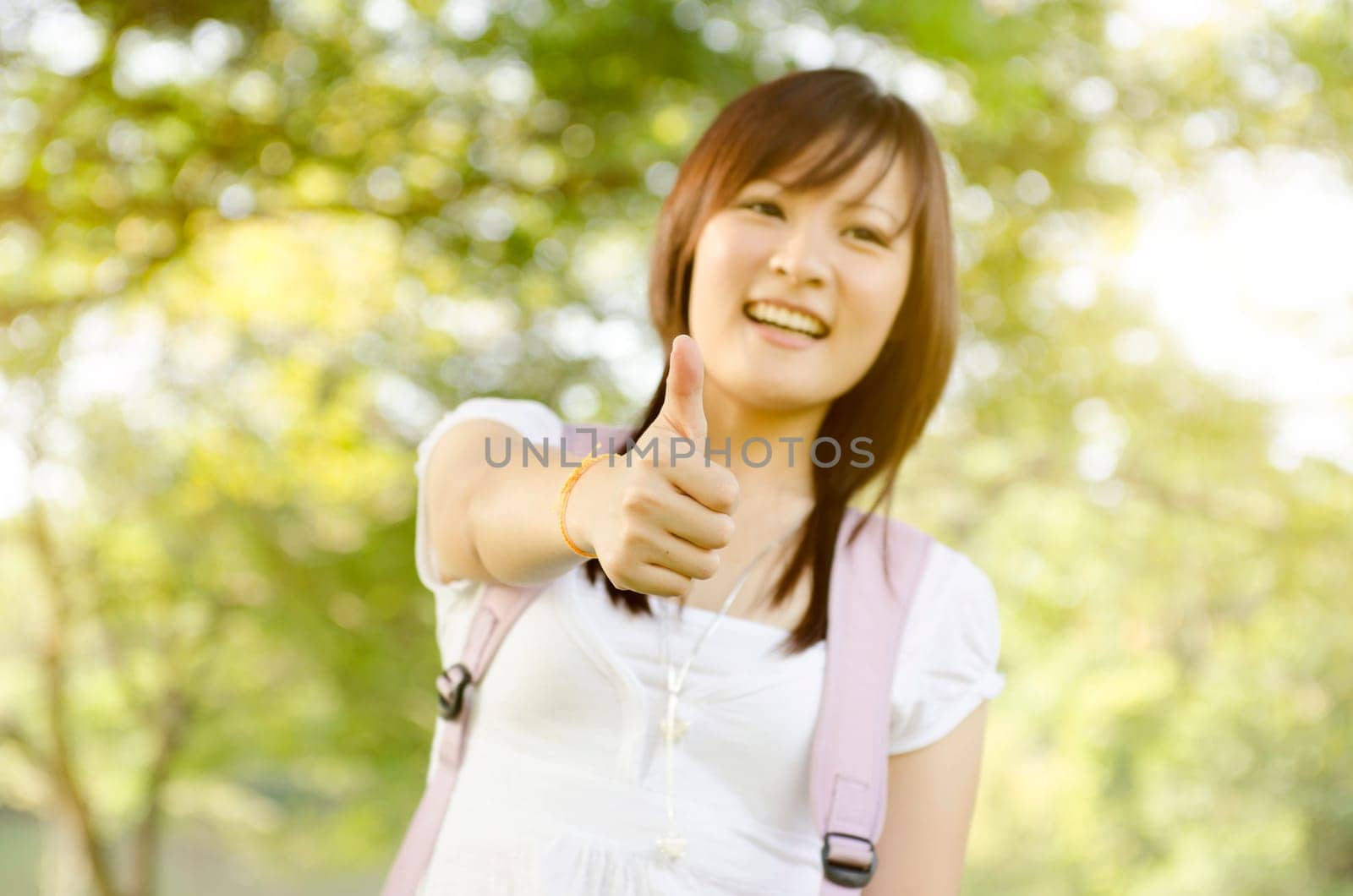 Young Asian college girl student standing on campus lawn, giving thumb up and smiling.