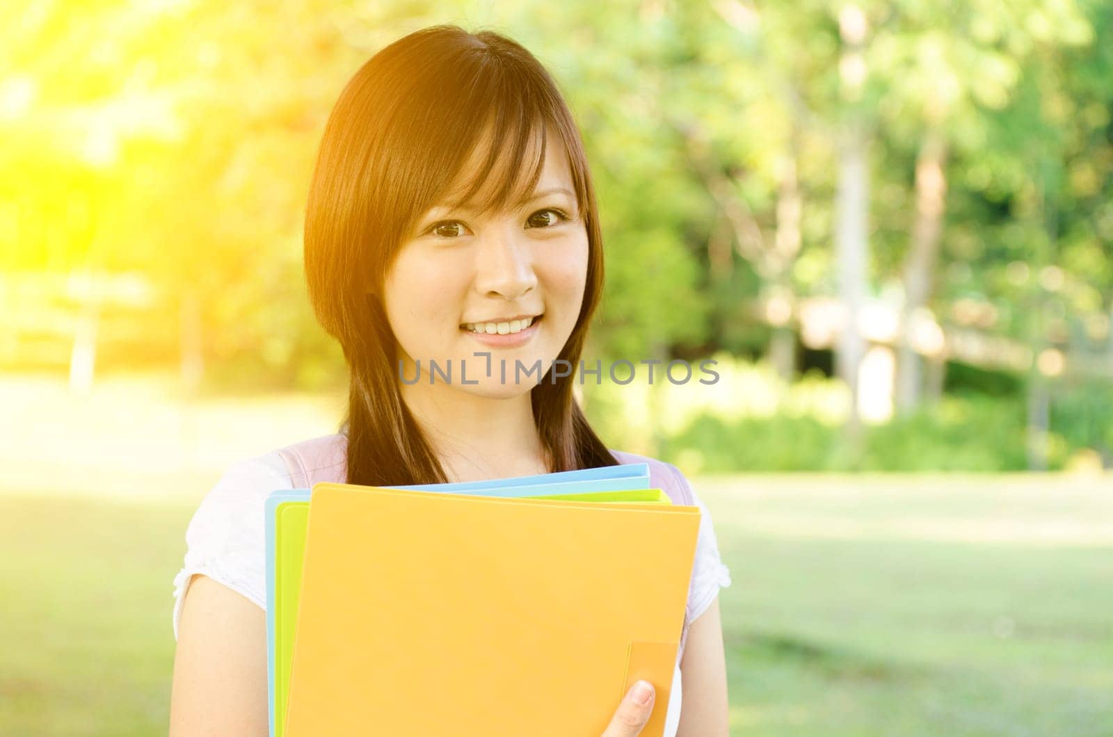 Young Asian college student standing on campus lawn, holding file folder and smiling.