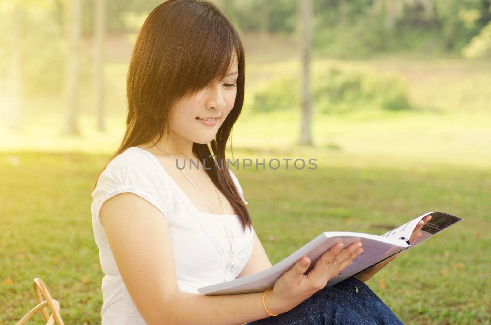 Young Asian college girl student sitting on campus lawn, reading book and smiling.