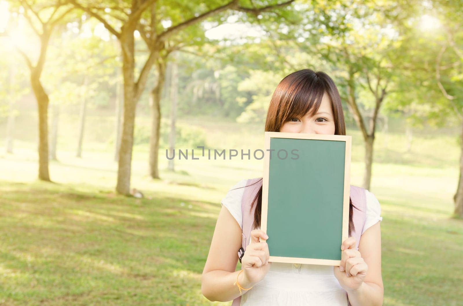 Young Asian college girl student standing on campus lawn, hands holding a blank chalkboard and covered mouth.