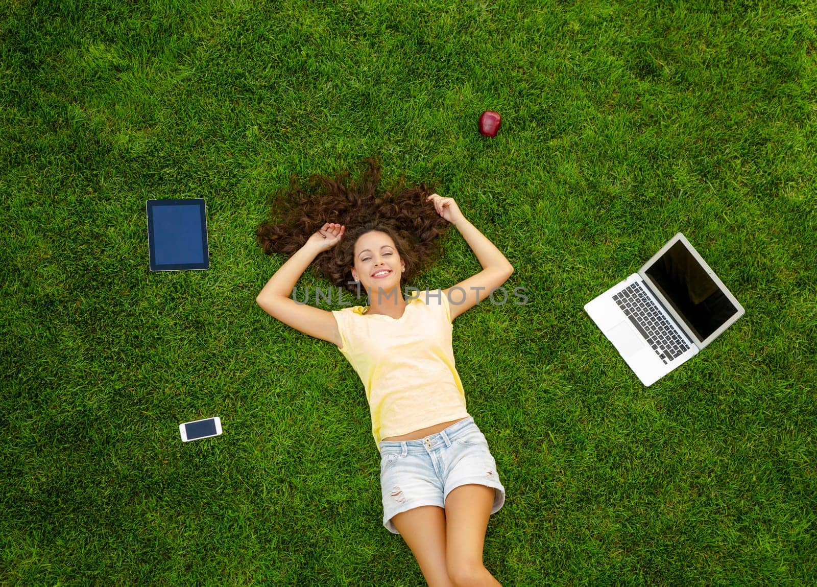 Beautiful and happy young woman lying on the grass surrounded by technology devices