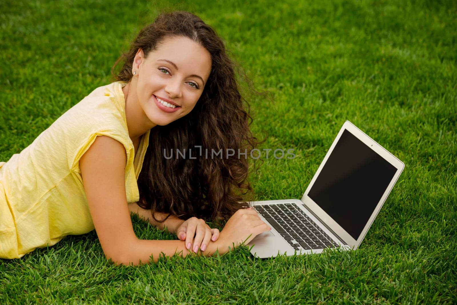 Beautiful young woman lying on the grass and working with a laptop