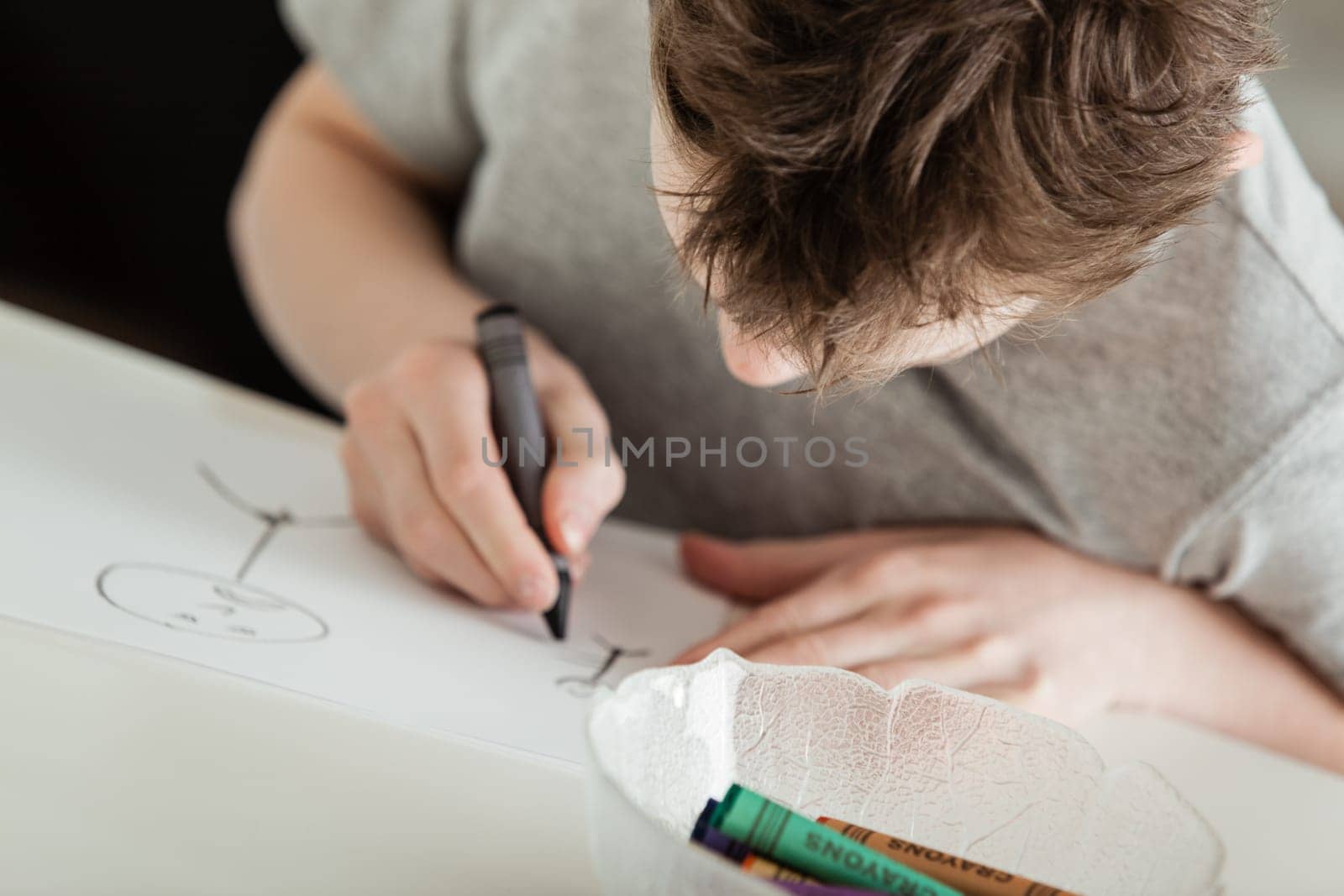 Serious Young Boy Draw Something on a White Paper Using Crayons at the Table.