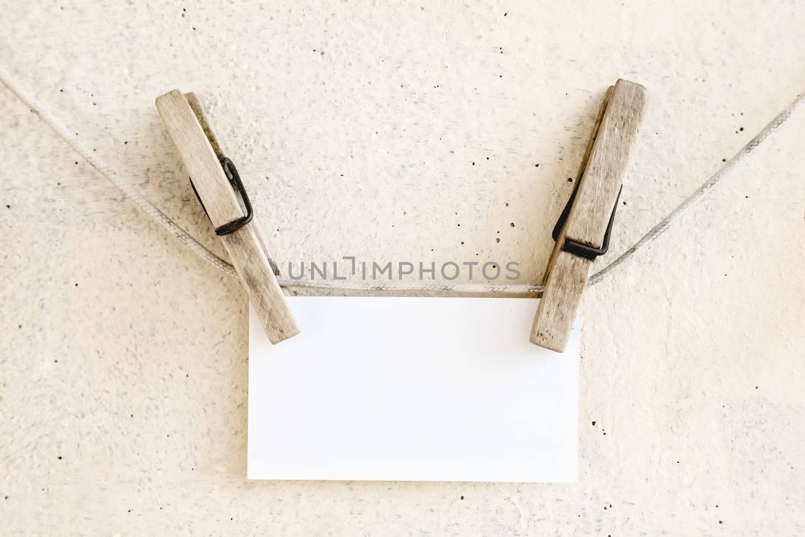 Clothespins holding on clothesline or rope holding white blank paper card on plaster background