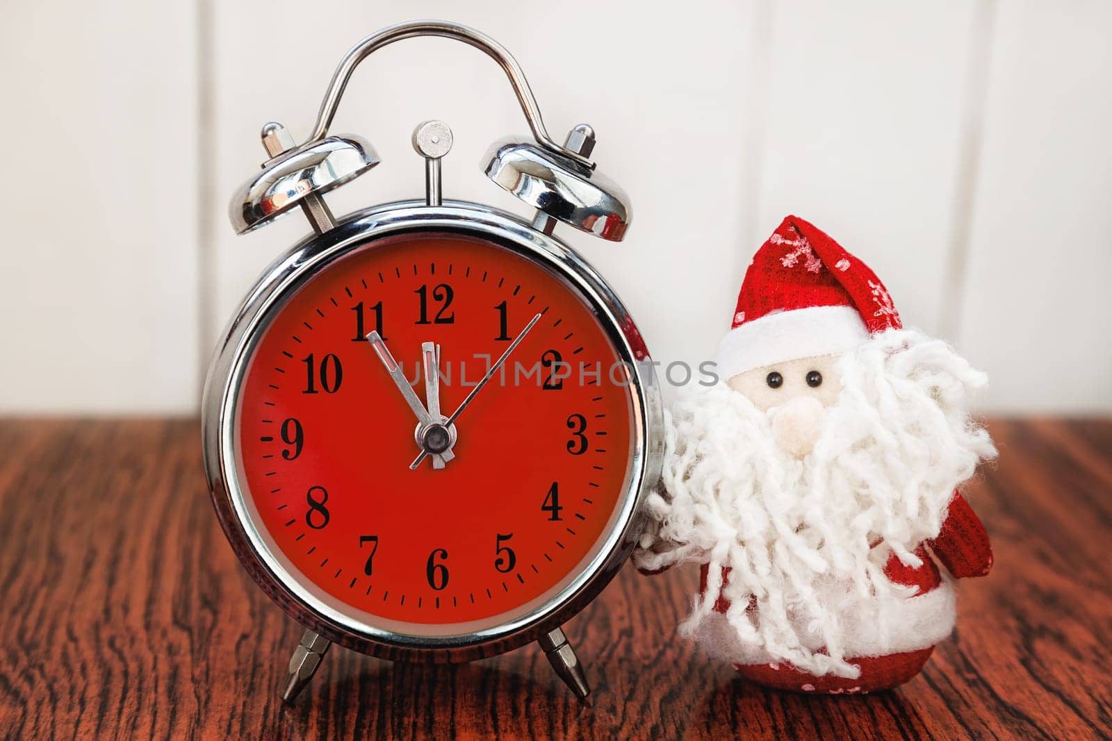Santa Claus or Father Frost and vintage alarm clock with red dial on wooden background. Showing time five minutes before twelve midnight