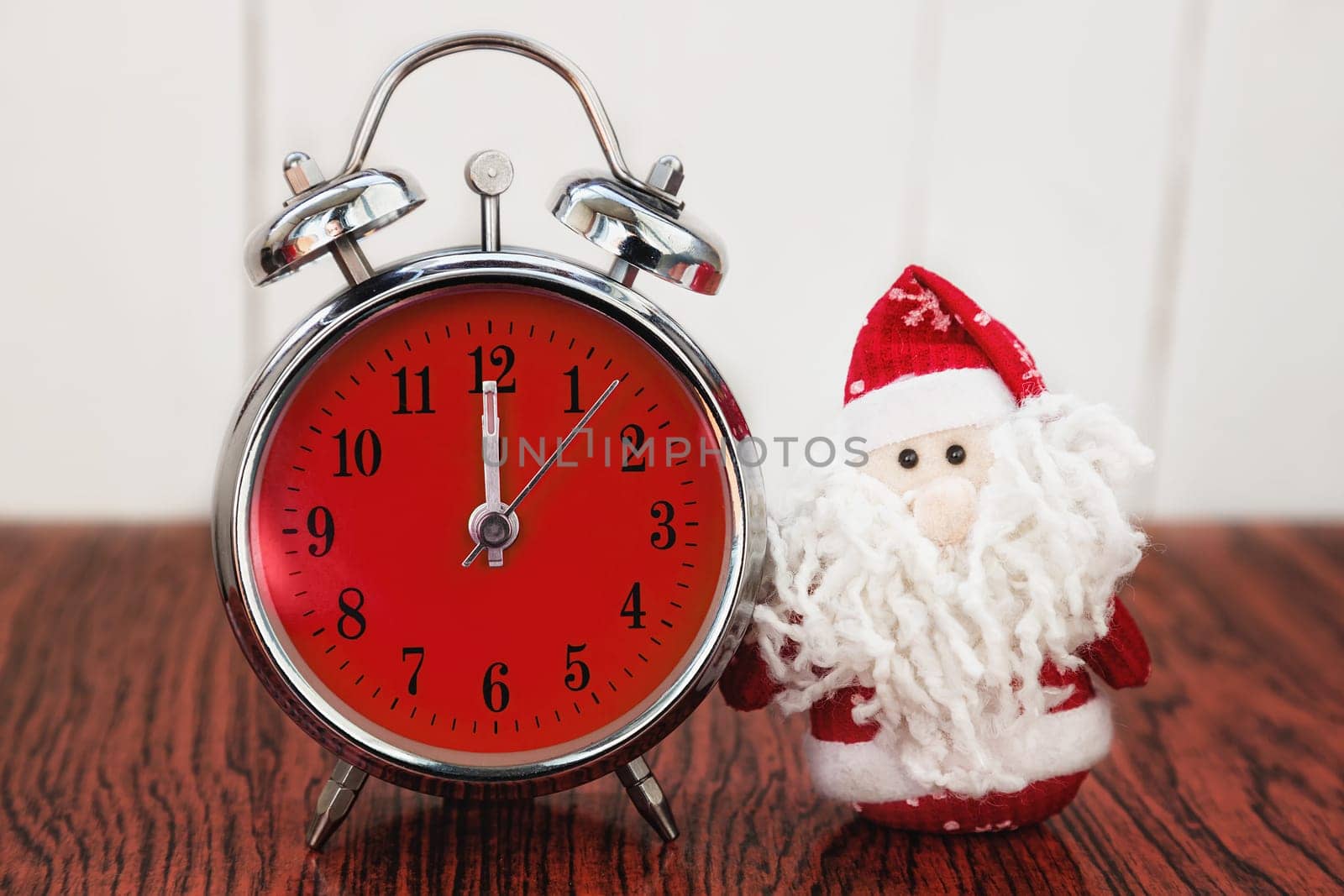 Santa Claus or Father Frost and vintage alarm clock with red dial on wooden background. Showing time twelve midnight