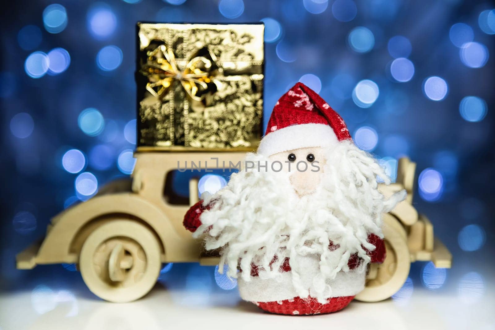 Santa Claus and old vintage wooden automobile with gift box on christmas lights background. Main focus of image on Santa Claus and defocused on side turned wood car with gift box