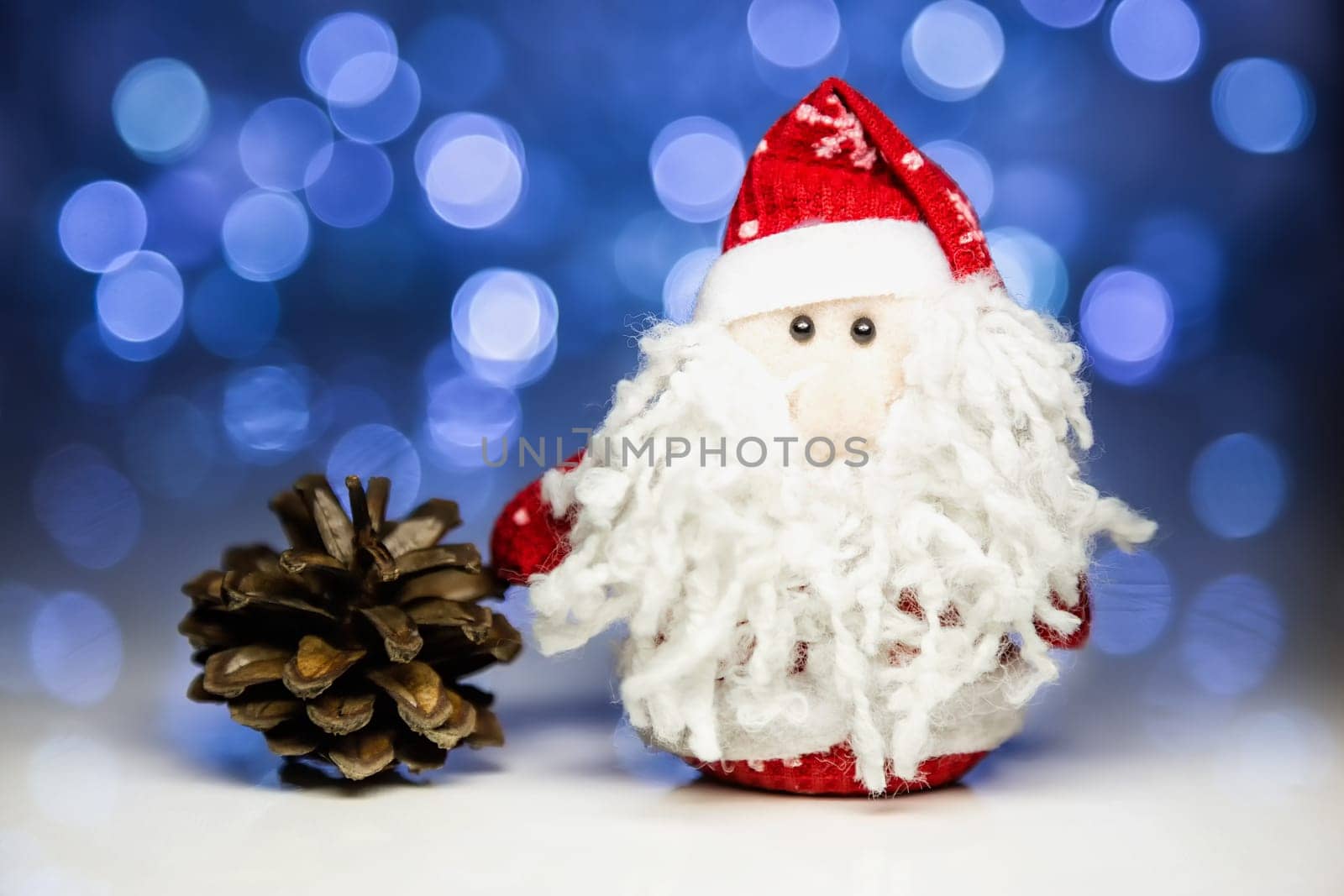 Santa Claus or Father Frost and pine cone with Christmas lights at background on white surface. Main focus of image on Santa Claus and selective on pine cone
