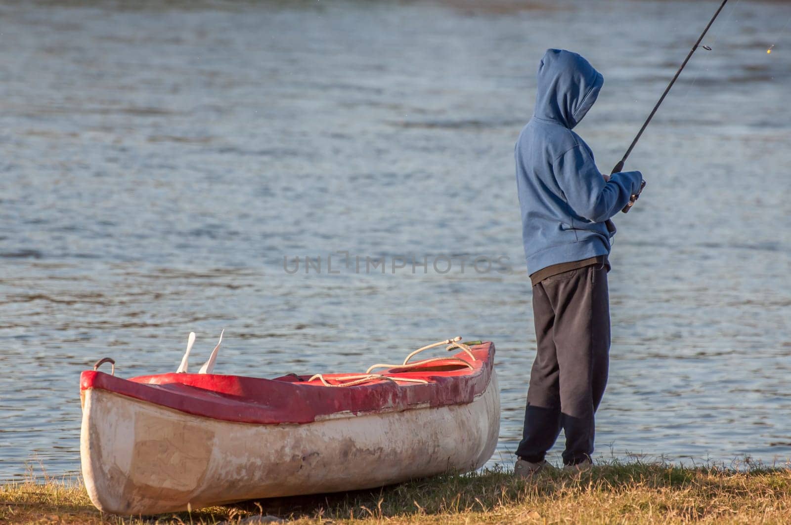Boy, standing next to canoe on the banks of a river, with fishing rod in hand, busy fishing.