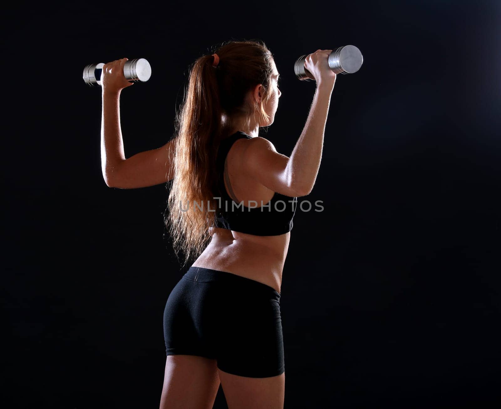 Young woman doing exercise with dumbbells, over black