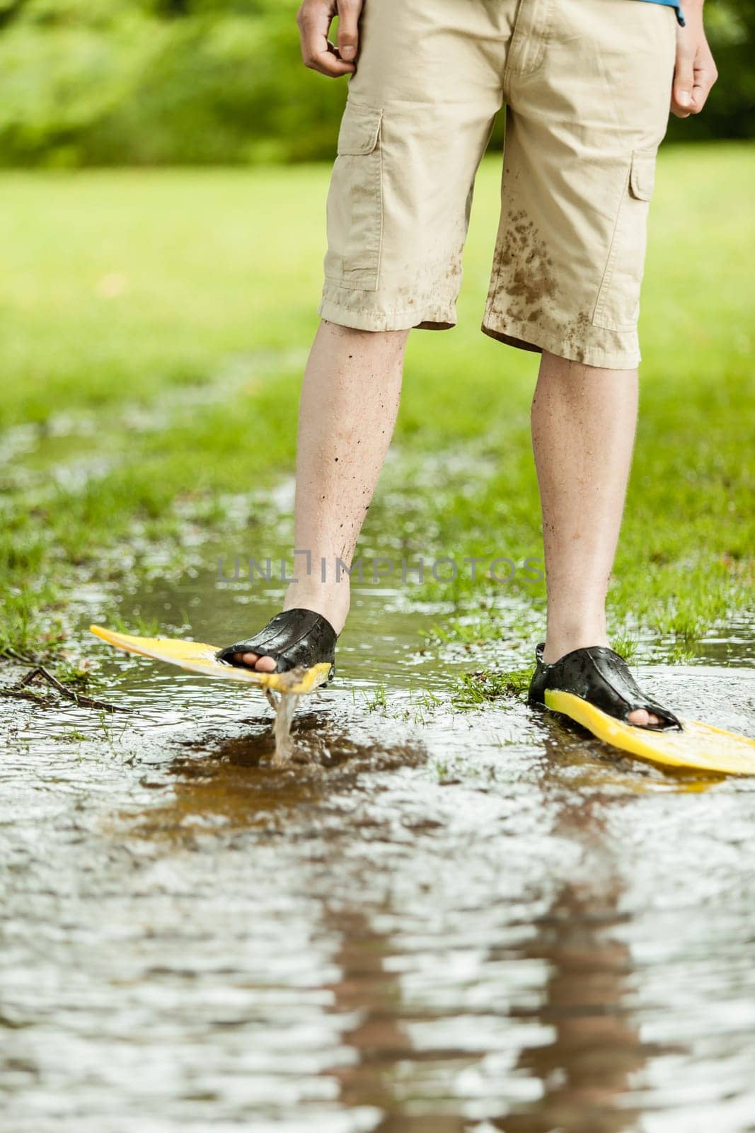 Unidentifiable person in shorts and diving flippers splashing in large puddle on turf grass