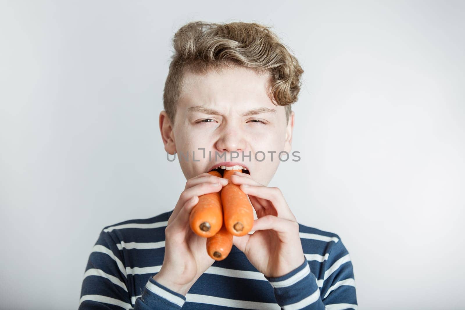 Head and Shoulders Portrait of Boy with Wavy Hair Stuffing Three Large Carrots into Mouth in front of White Studio Background with Copy Space - Health and Nutrition Humorous Image