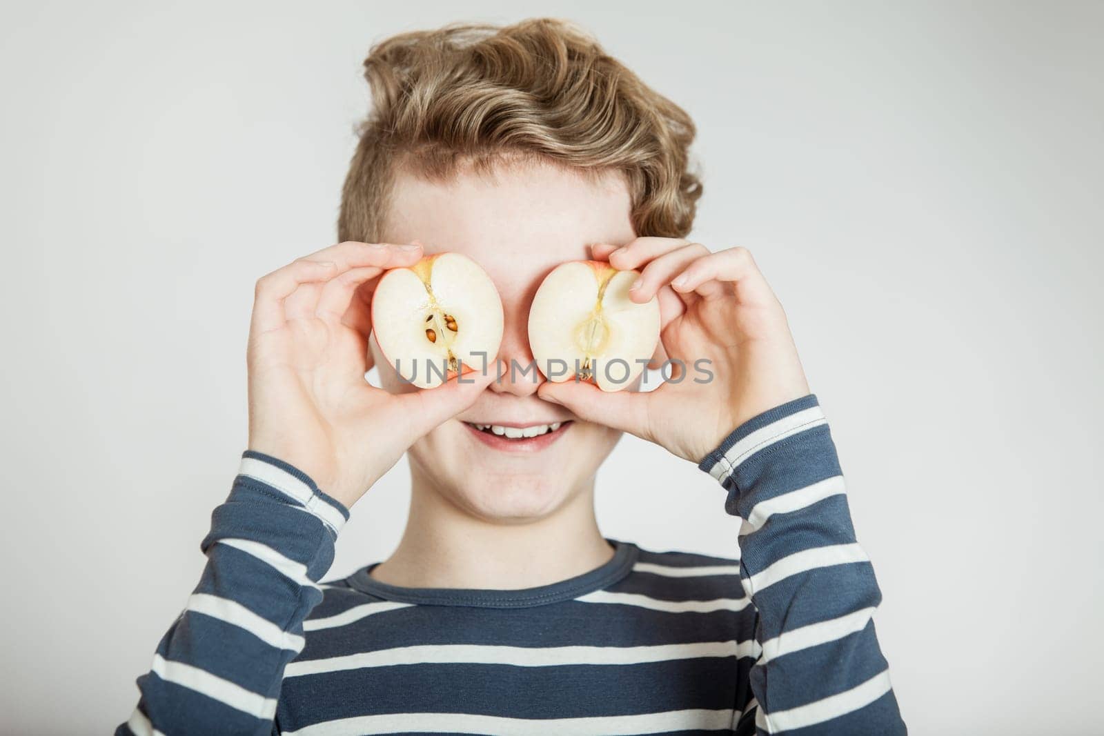 Giggling child in striped blue and white shirt holding apple halves in front of his eyes over gray background