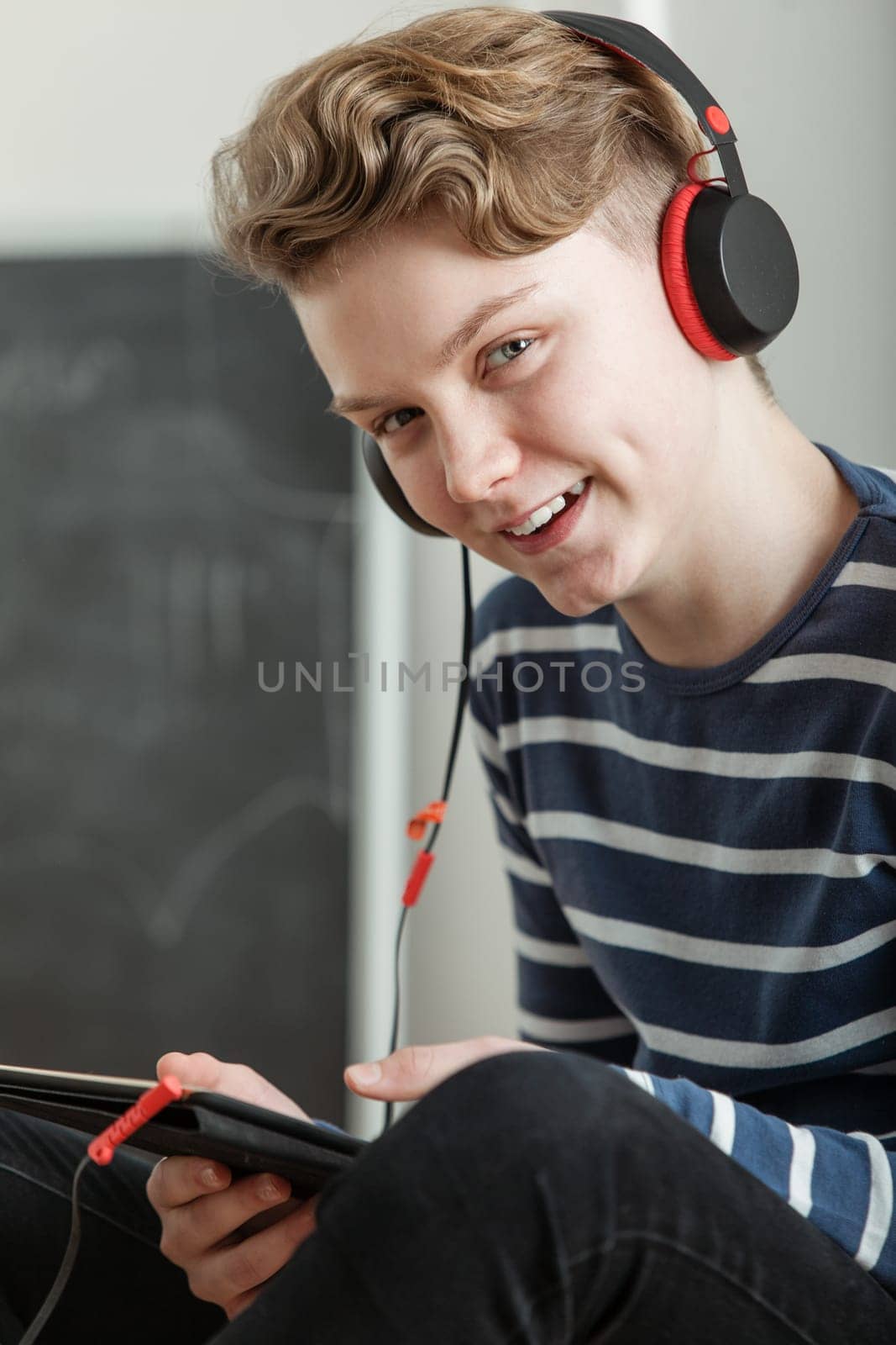 Waist Up Portrait of Teenage Boy with Wavy Blond Hair Listening to Music Through Headphones and Holding Handheld Tablet Computer in Classroom, Smiling at Camera