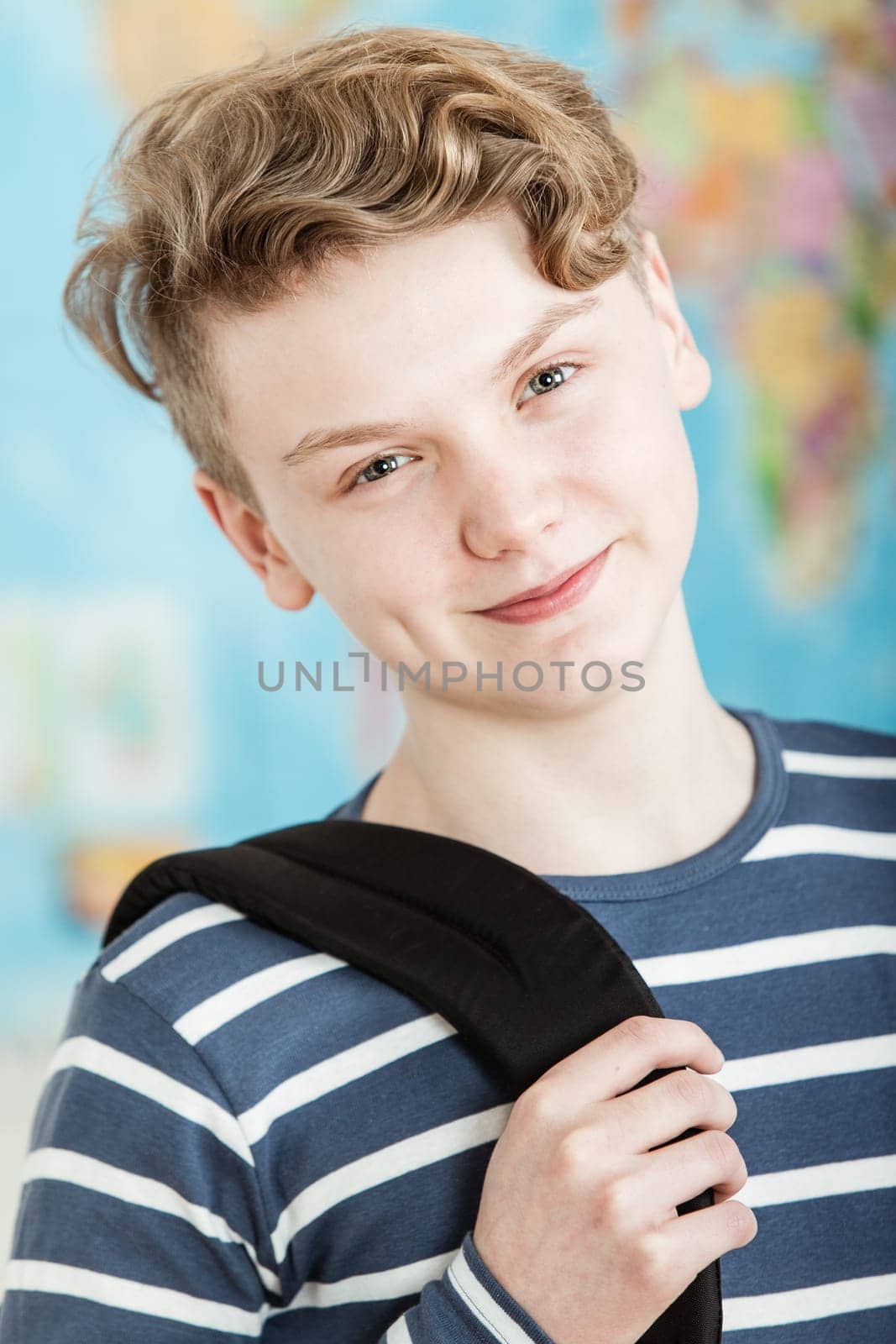 Head and Shoulders Portrait of Smiling Boy with Wavy Blond Hair Standing in Classroom with World Map in Background, Wearing Blue and White Striped Shirt and Holding Knapsack
