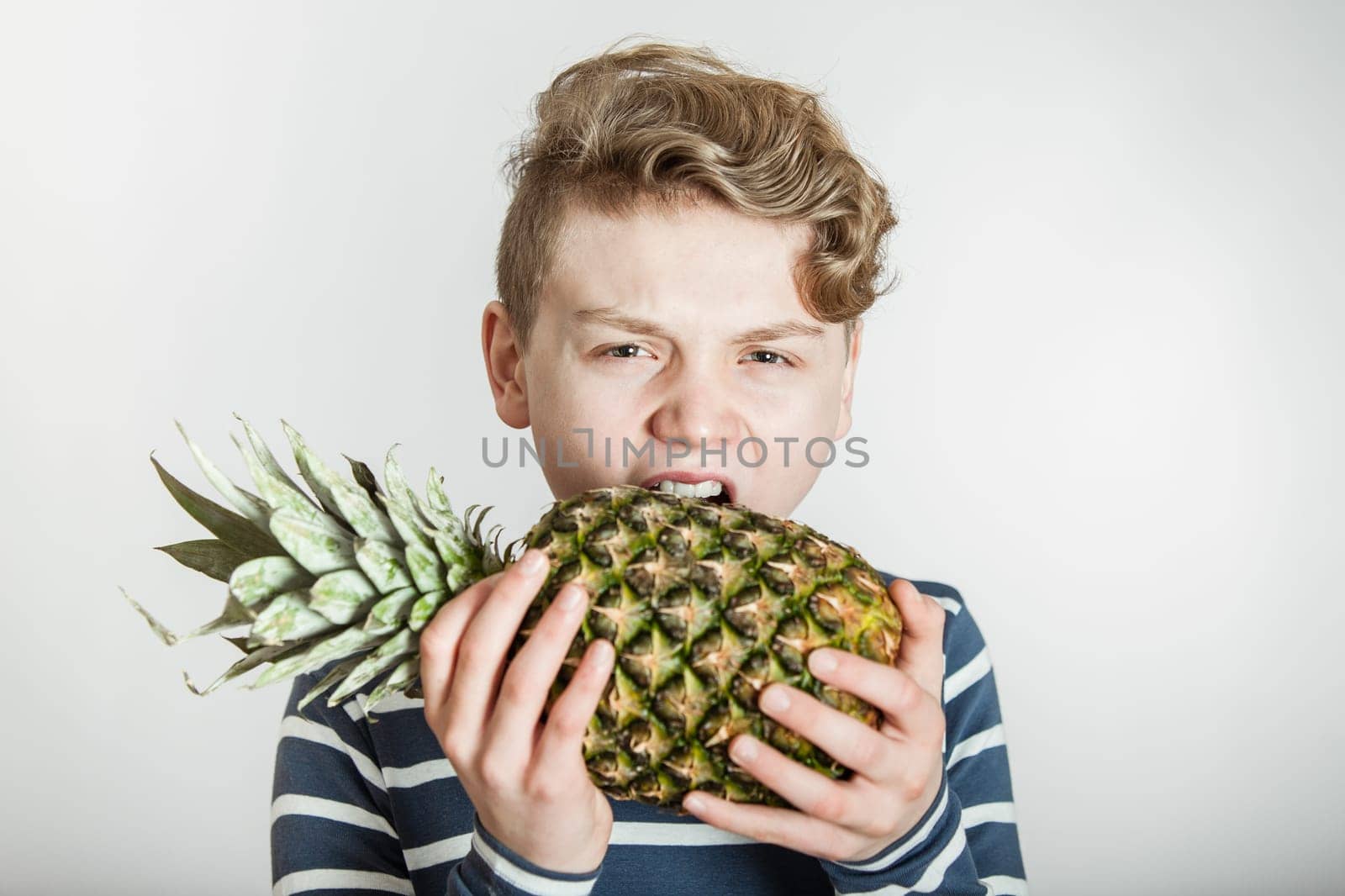 Head and Shoulders of Boy with Wavy Blond Hair Biting into Tough Skin of Spikey Pineapple in Studio with White Background with Copy Space