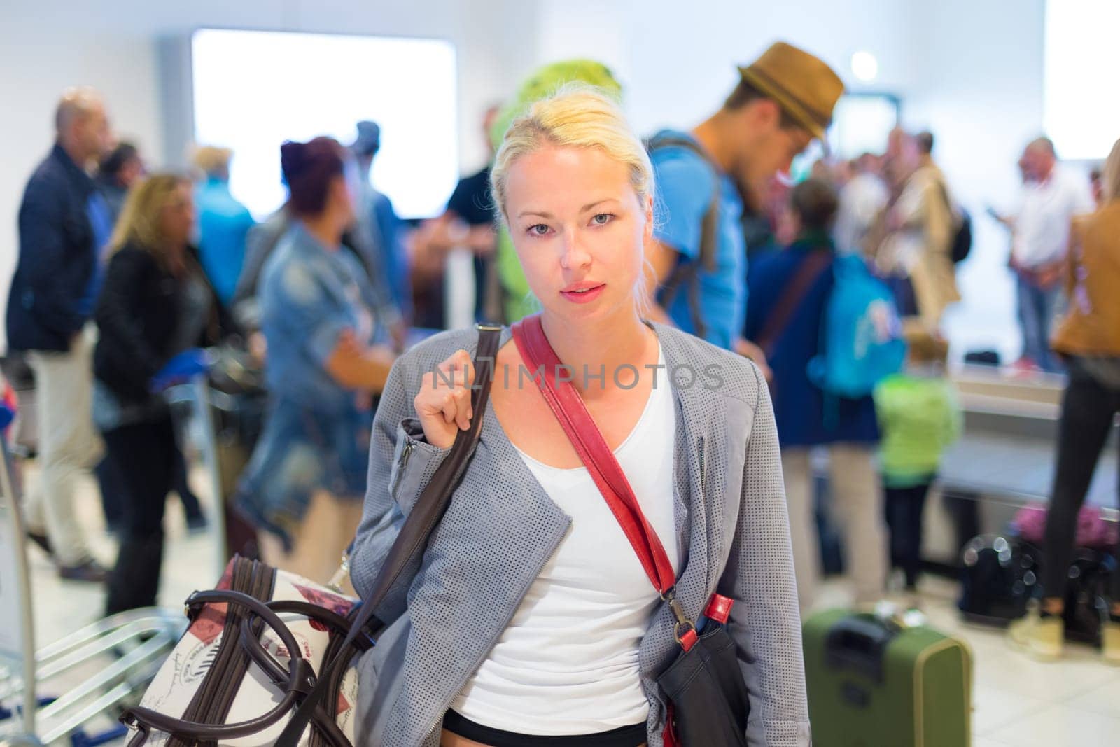 Casually dressed young stylish female traveller waiting in airport arrival terminal hall,  collecting her laggage . Blured background. Can also be used as railway, metro, bus station.