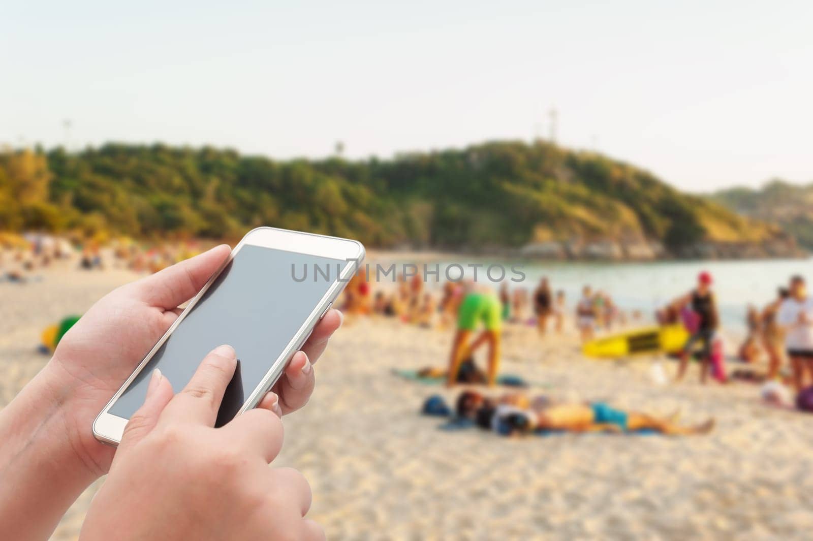 Women using smartphone top on blurred sea and sand beach with some people in evening light - Can be used for display or montage your products