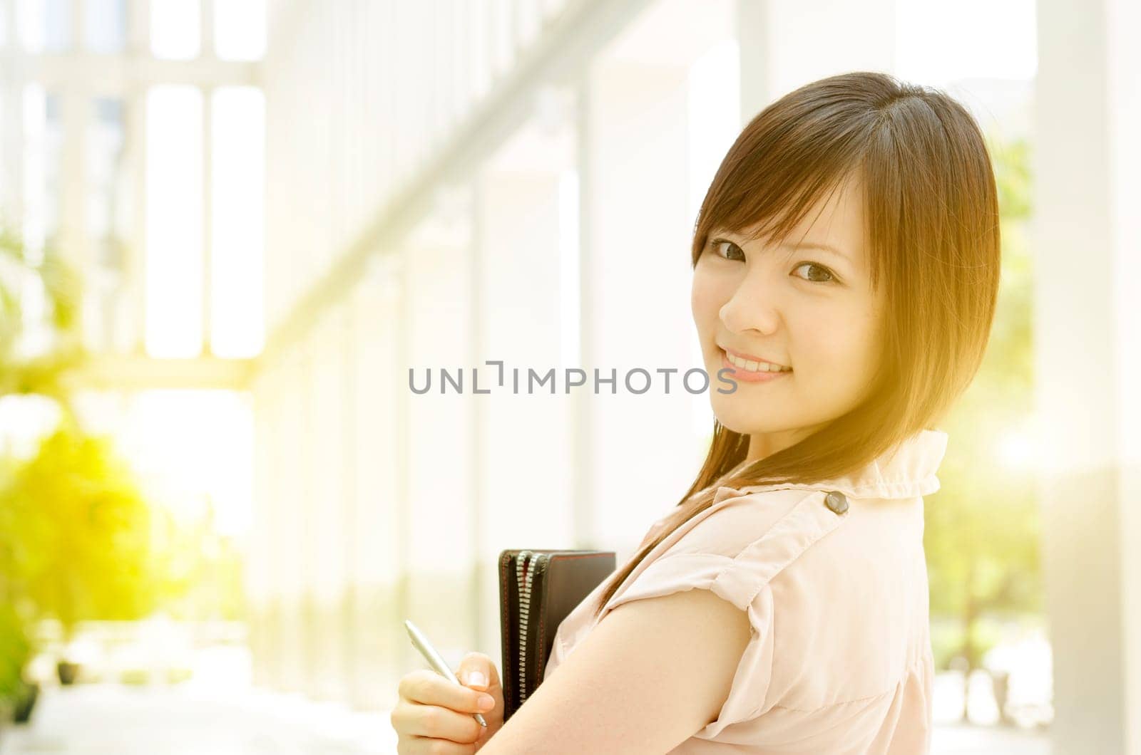 Young Asian female business people smiling and holding file folder, standing in an office environment, beautiful golden sunlight at background.