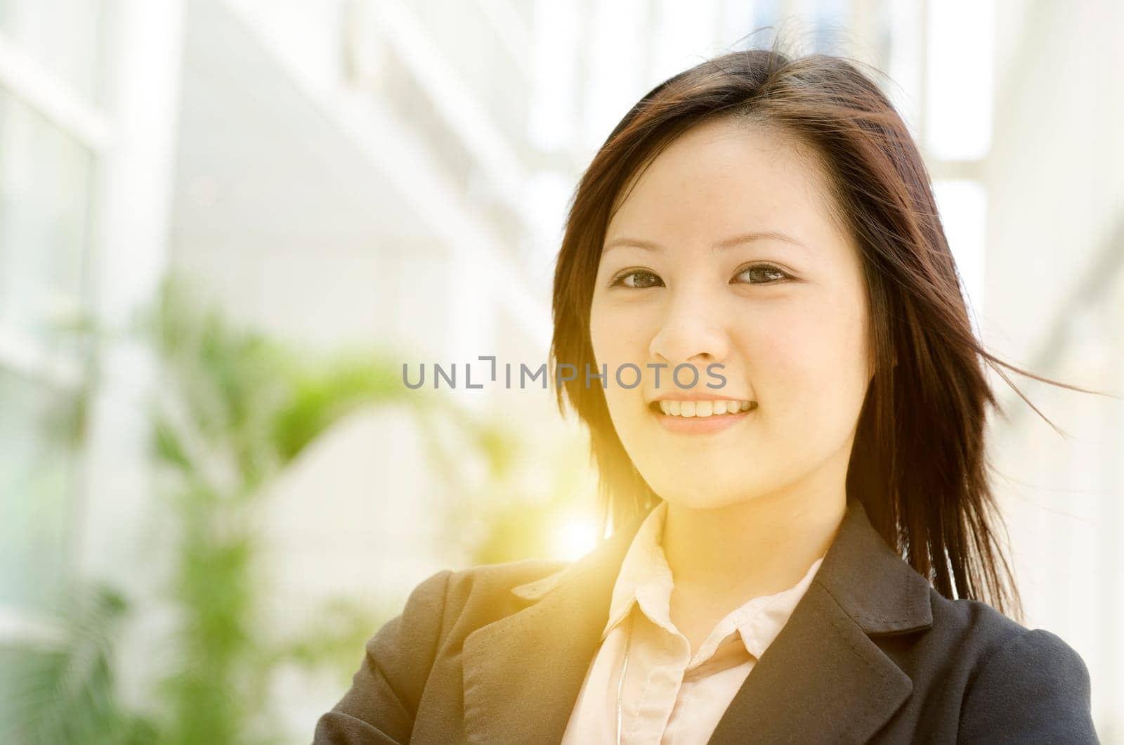 Portrait of a young Asian business woman smiling and standing at an office environment, natural golden sunlight at background.