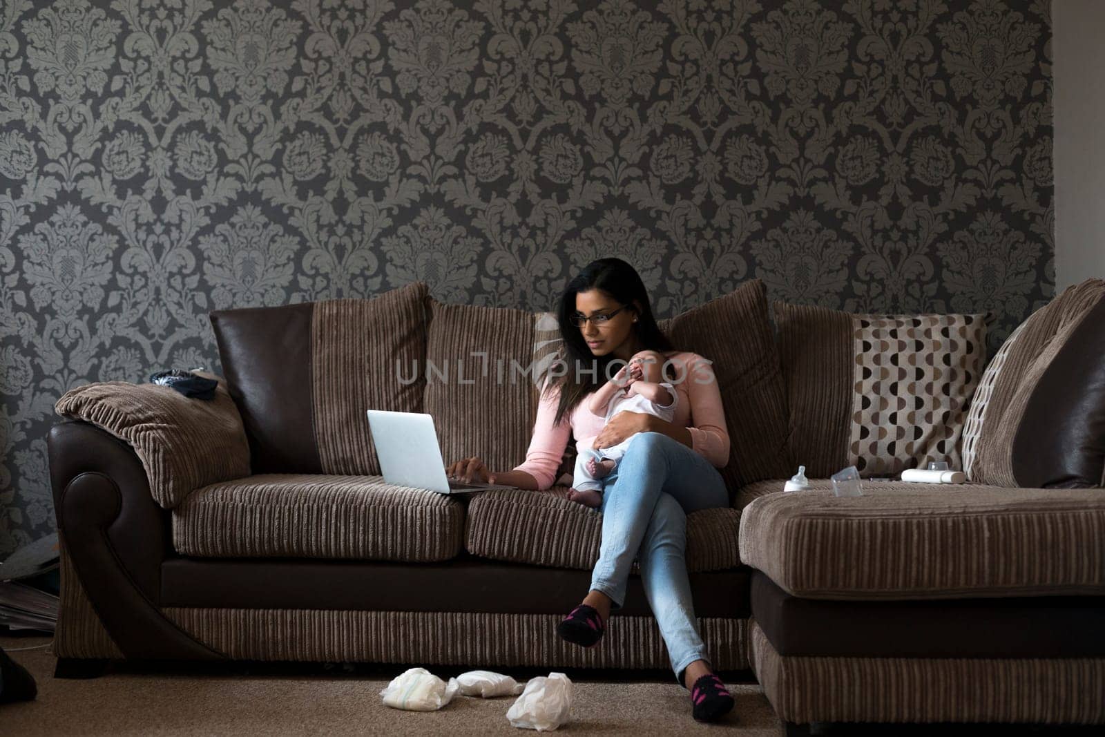 Young mother sitting on her sofa at home with her baby son on her knee. She is working on a laptop and there are dirty nappies on the floor.