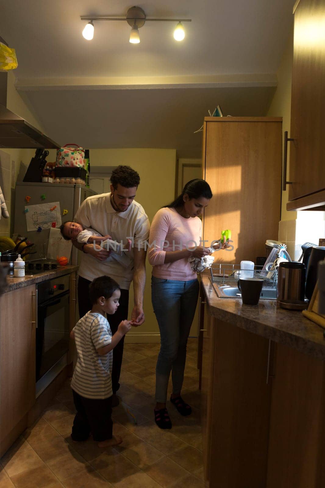 Young family in the kitchen. The mother is washing dishes while the father is holding their baby son and tending to his eldest who has hurt his finger.