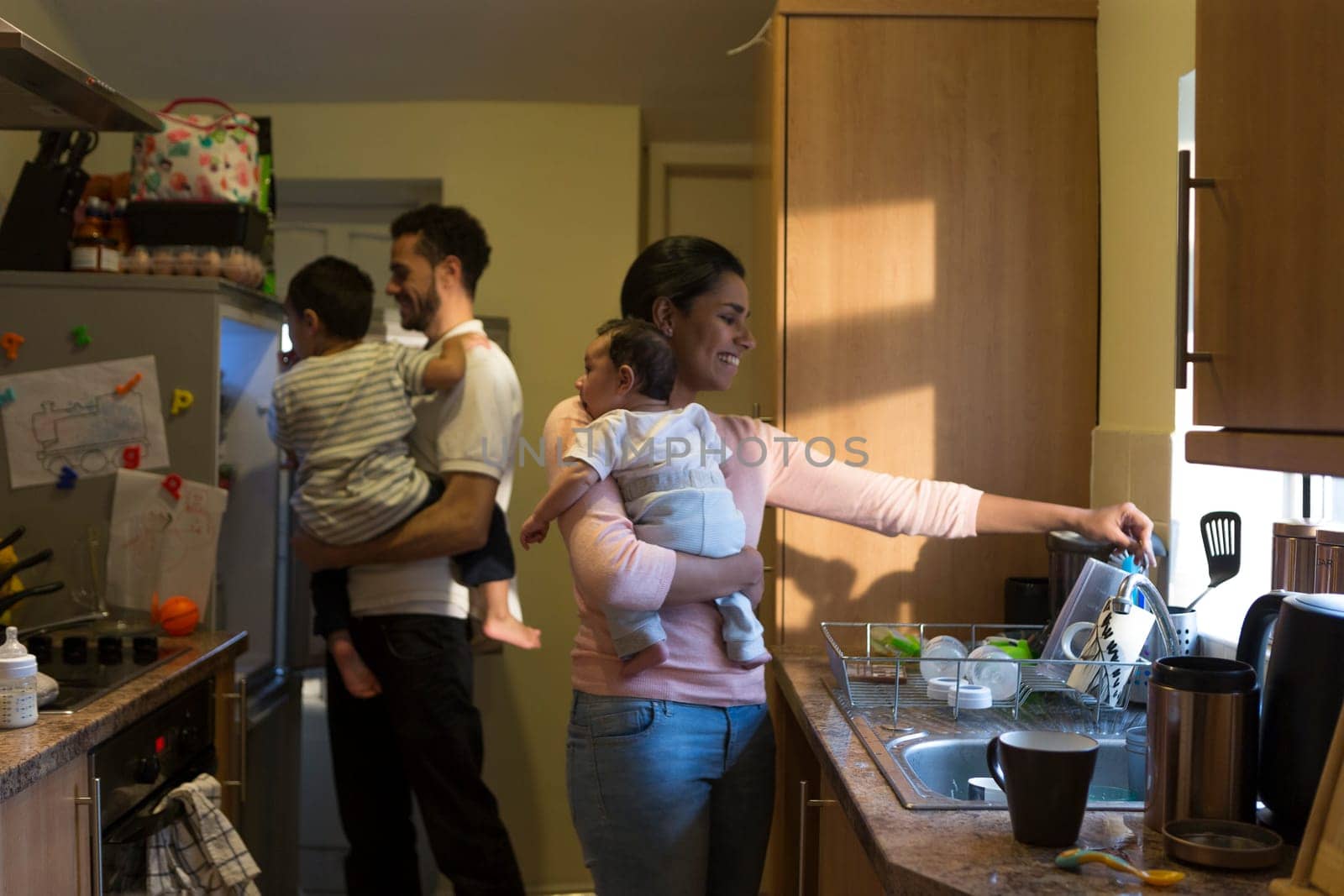 Young family in the kitchen. The mother is washing dishes with their baby on her shoulder while the father is looking in the fridge with their eldest on his shoulder.