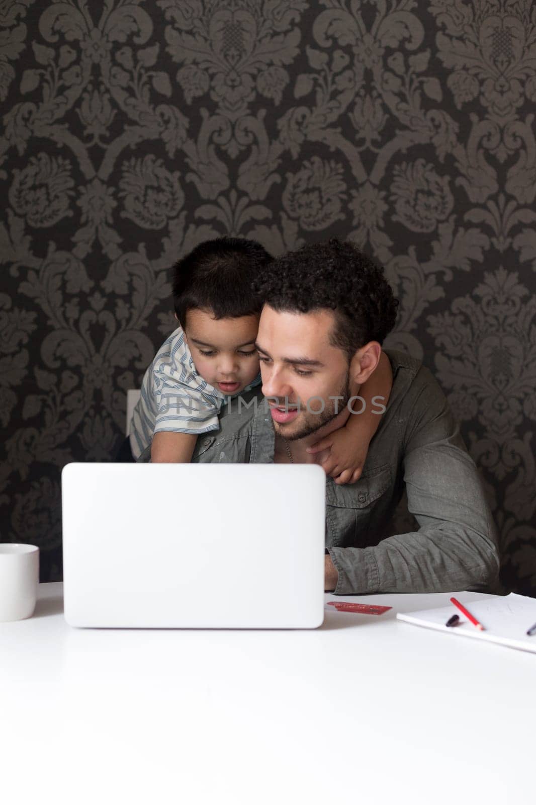 Little boy is climbing on his dad, trying to help him with his internet banking. They are sitting behind a laptop at home.