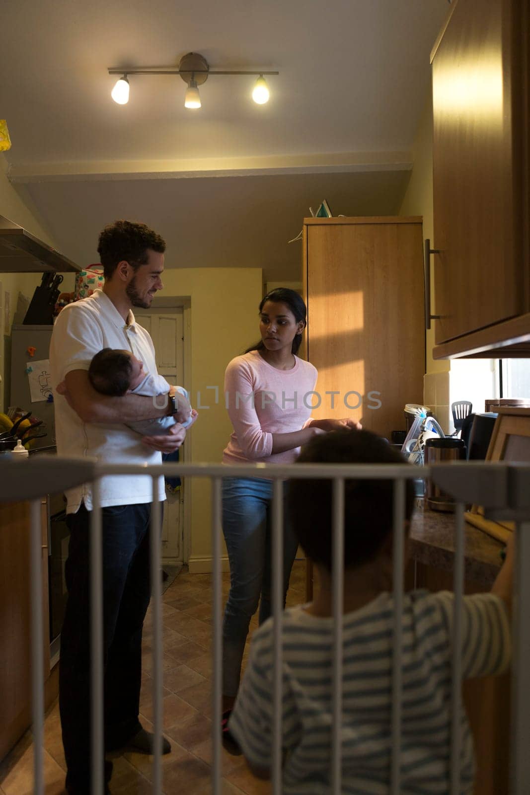 Young family in the kitchen. The mother is washing dishes and talking to her partner, who is holding their baby son, The eldest son is standing with his back to the camera, playing with his toy car on the worktop.