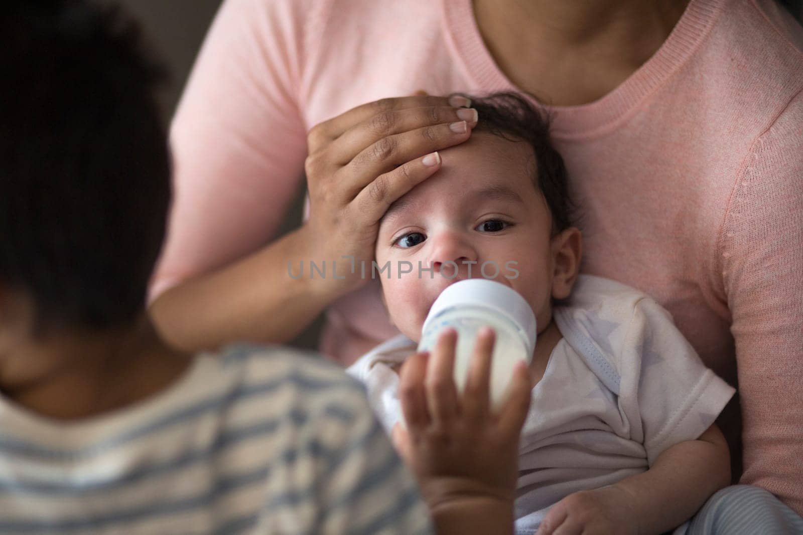 Close up shot of a baby boy being fed a bottle of milk by his older brother whilst sitting on his mother's lap.