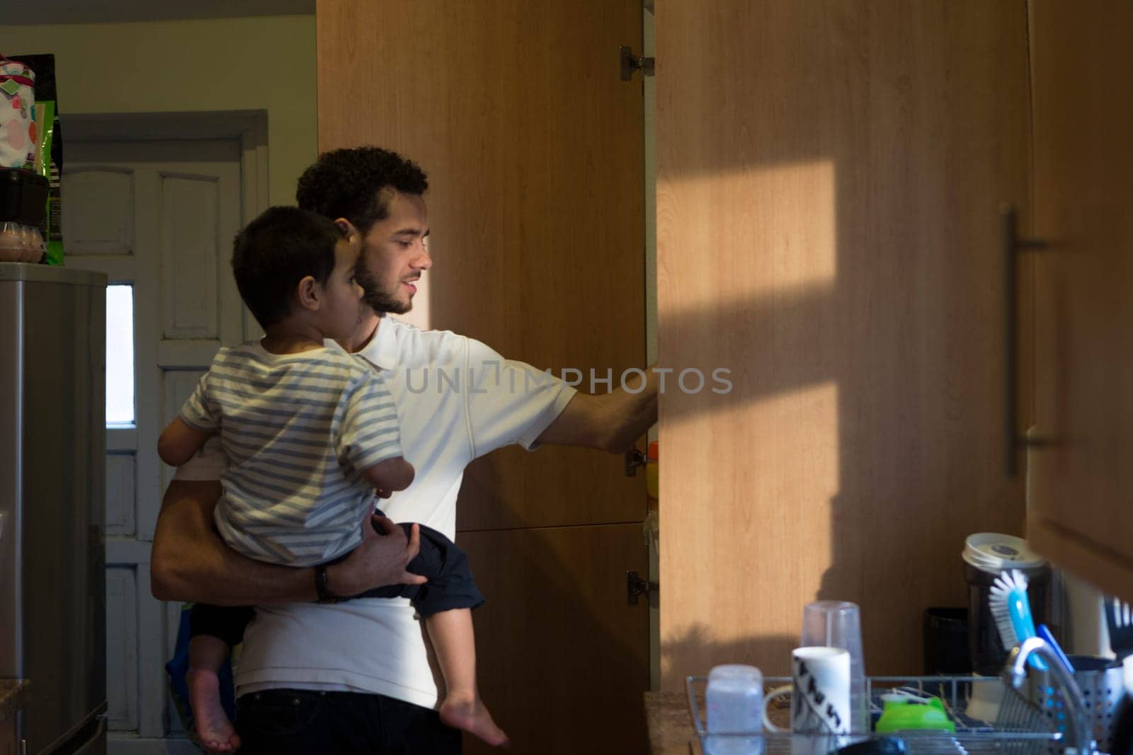 Young father in the kitchen, getting something out of the cupboard with one hand with his son on his shoulder.