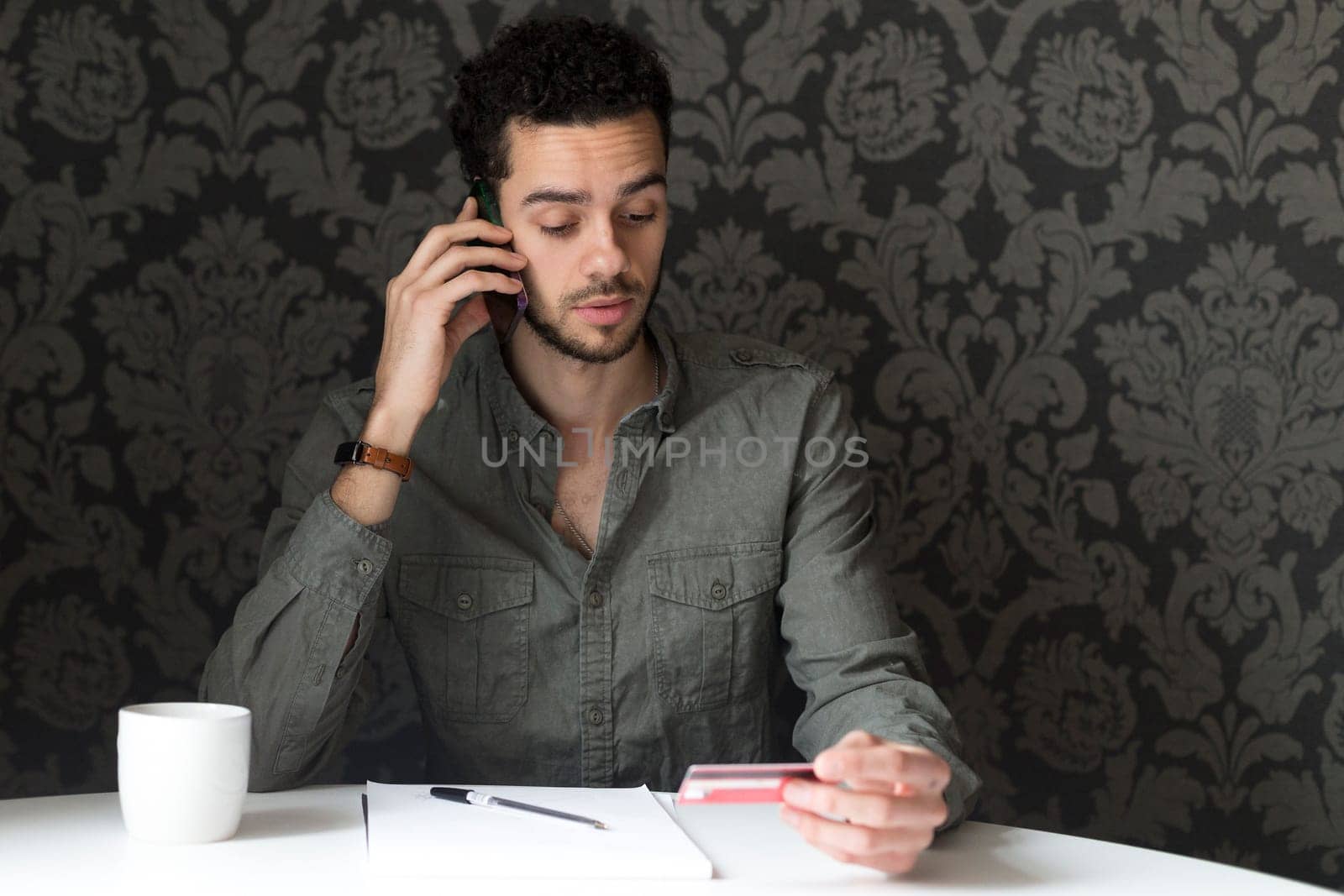 Young man sitting at the table at home, using his smartphone and his bank card to pay bills over the phone.