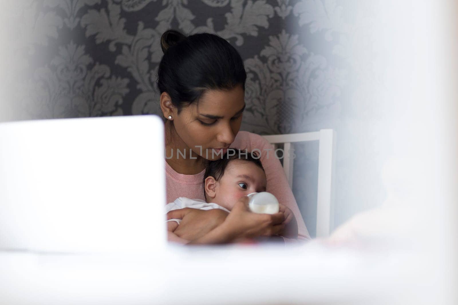 Young mother feeding her baby son a bottle of milk whilst sitting at her computer.