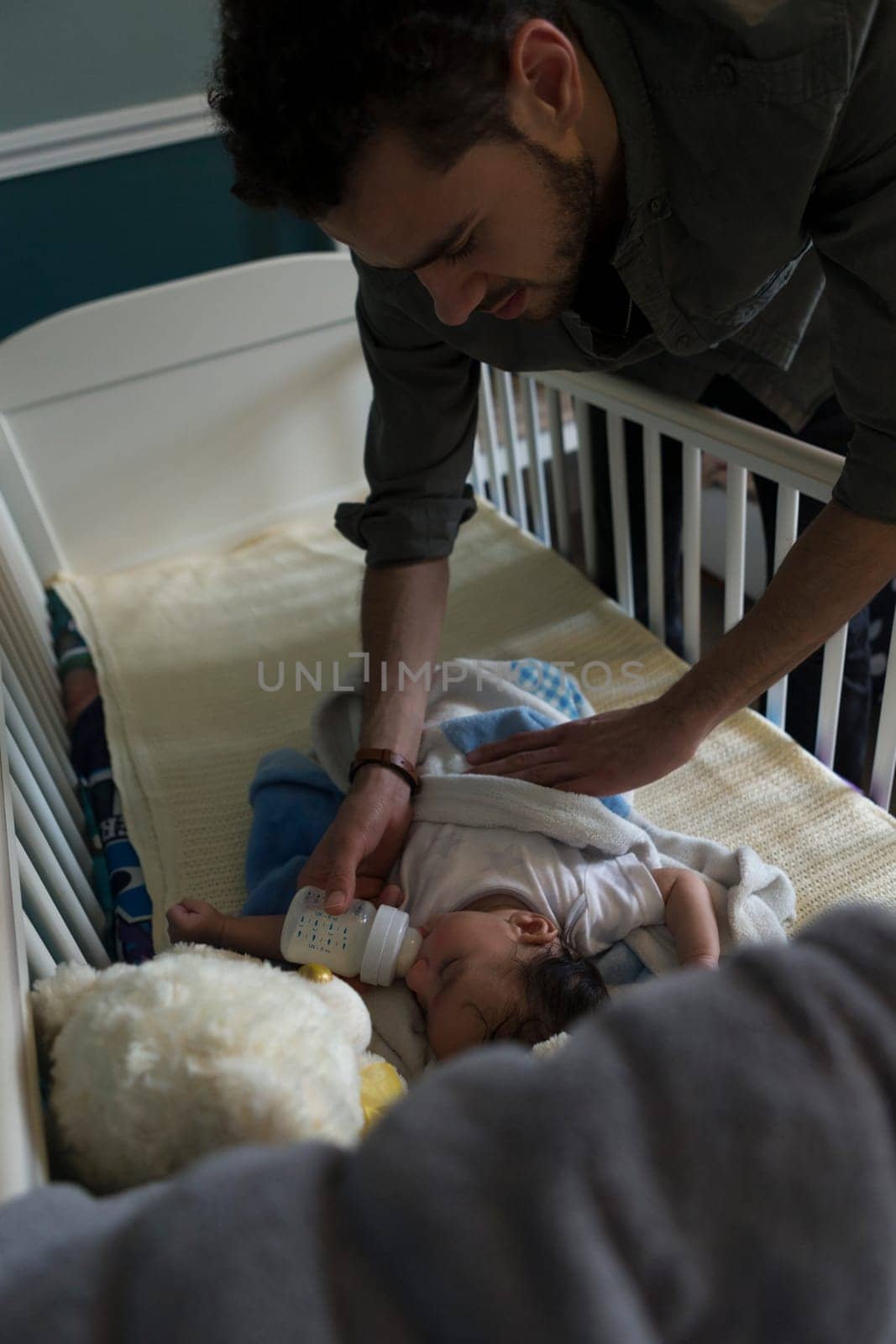 Young father feeding his son a bottle. He is lying in his cot, nodding off to sleep.