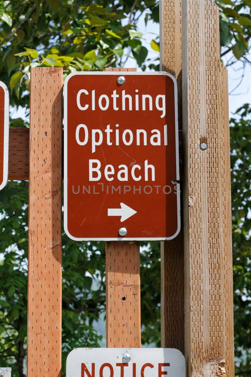 On a wooden post is a brown and white metal Clothing Optional Beach sign on wooden posts outside with foliage in the background on a sunny day.