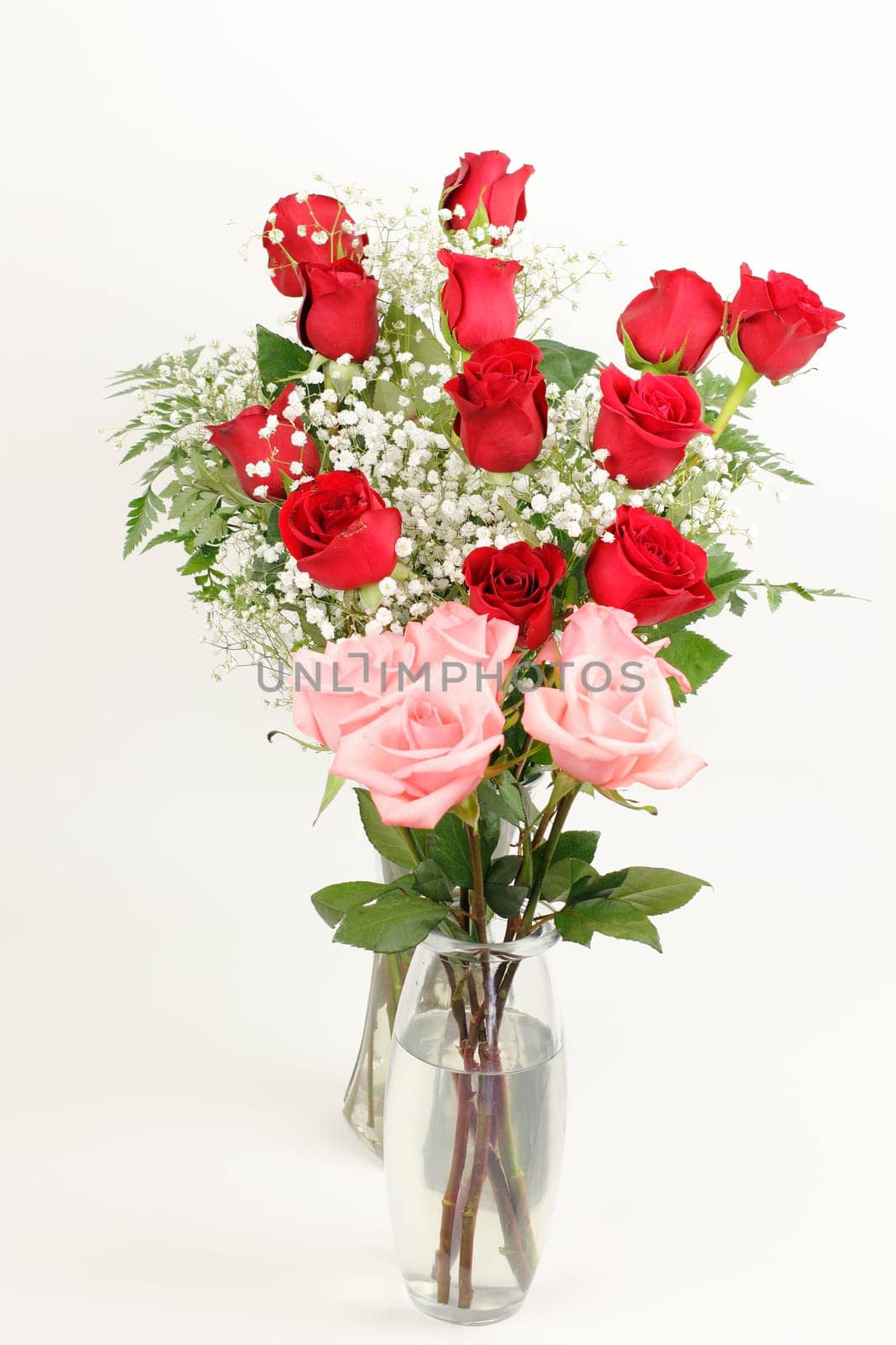 One glass vase bouquet of pink roses  in front of a glass vase of red rose s floral arrangement in front of an off white background. Two pretty rose bouquets in glass vases.