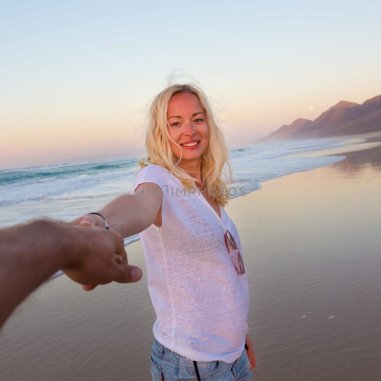 Young romantic couple, holding hands, having fun on perfect deserted beach at sunset. Shot from boyfrieds perspective. Guy looking at her beautiful carefree girlfriend.