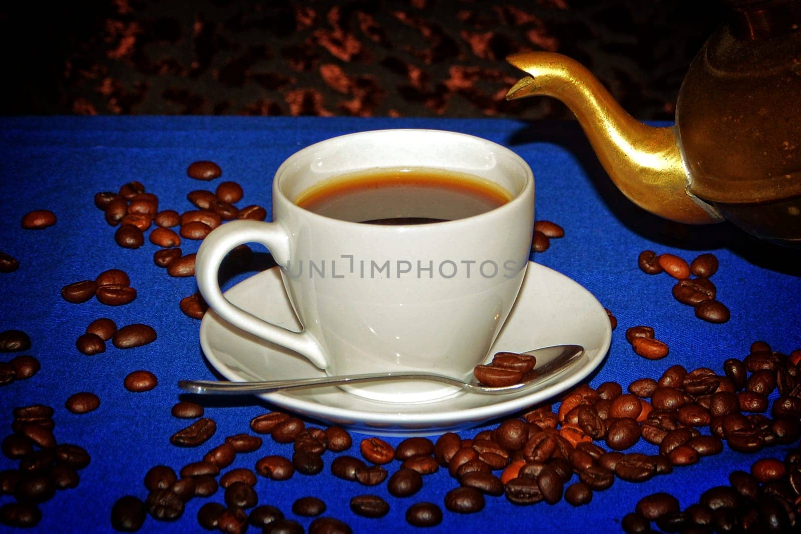 White cup of coffee on blue tablecloth with coffee beans and neck of a golden jug
