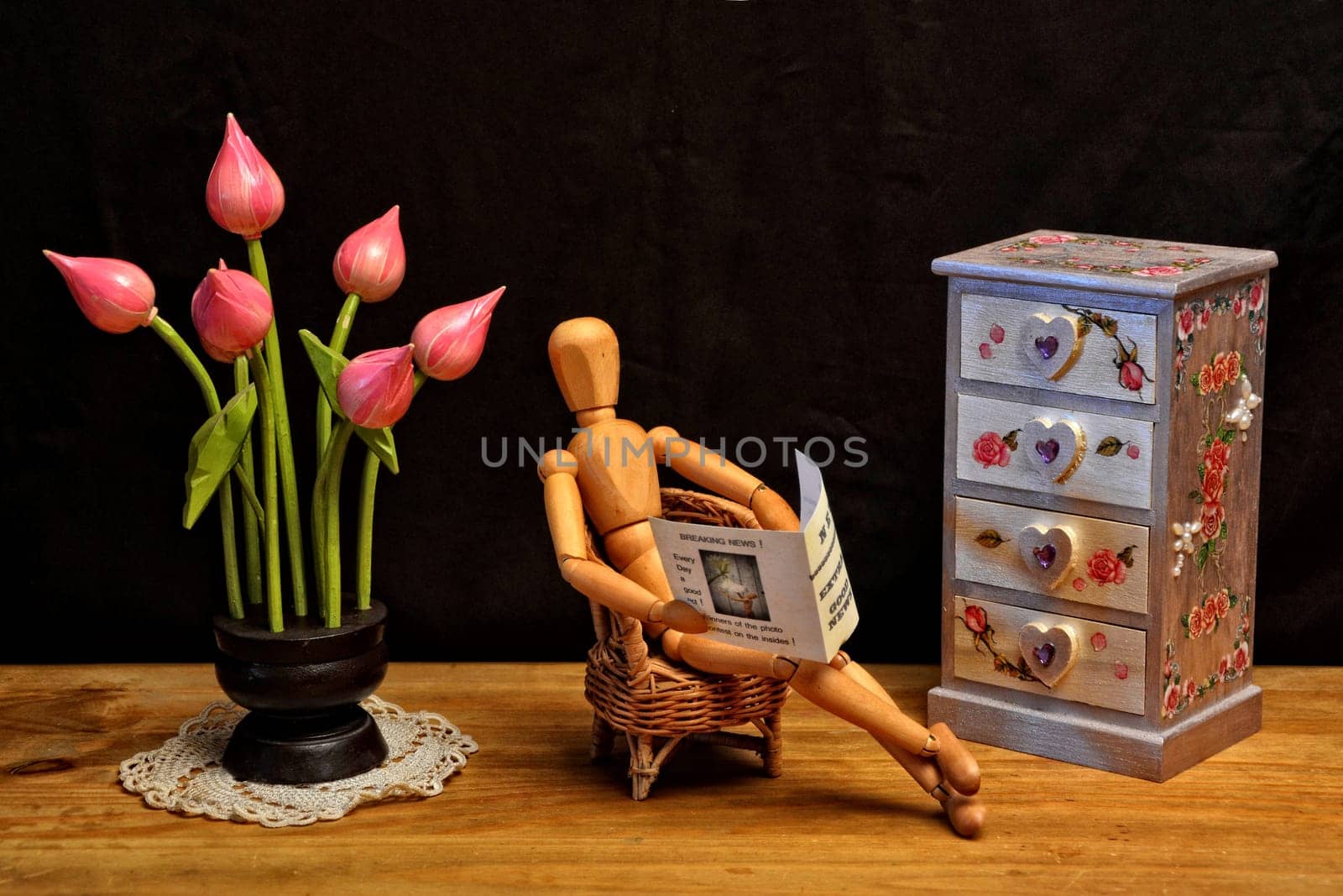 Wooden figure reading newspaper in the living room, next to dresser and wooden flowers with dark background