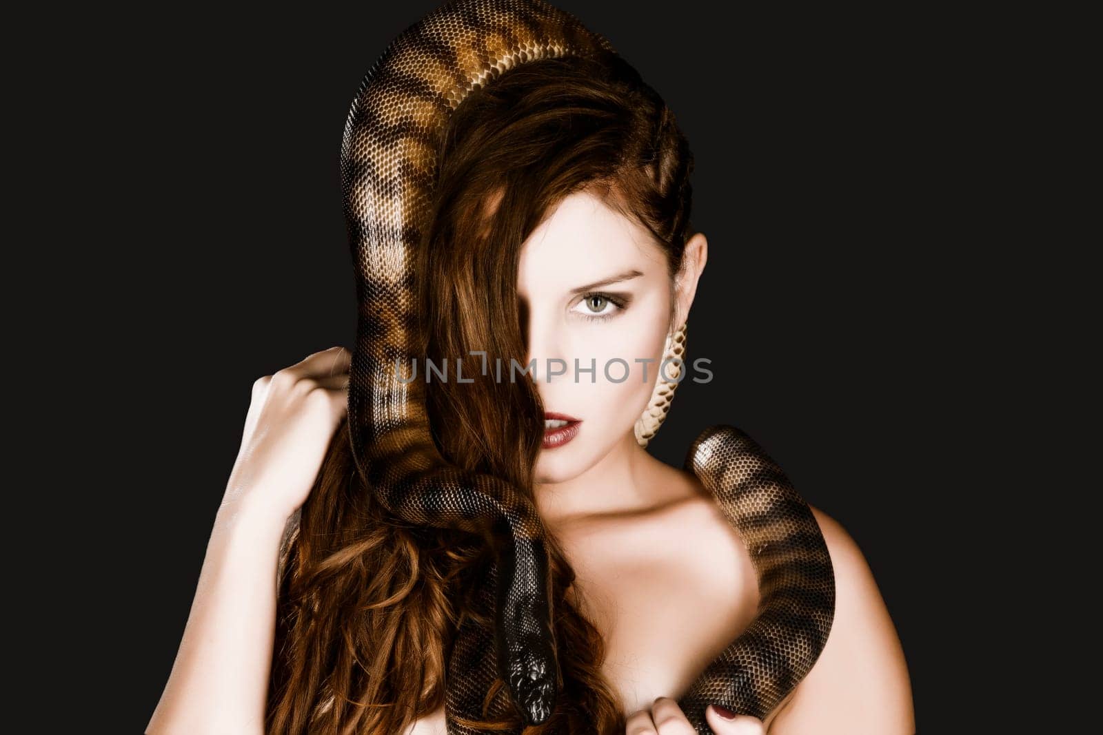 A pretty young woman with brunette hair looks at the camera head to her bare neck snaking a big dark snake.