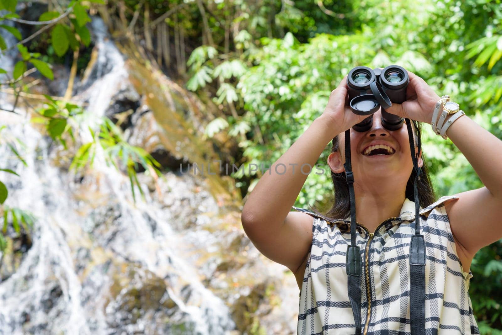 Beautiful young girl hiking is using binoculars look for birds in tropical forest near the waterfalls in Thailand