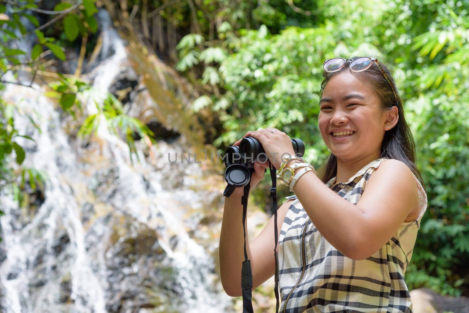 Beautiful young girl hiking is using binoculars look for birds smiling to the camera in tropical forest near the waterfalls in Thailand